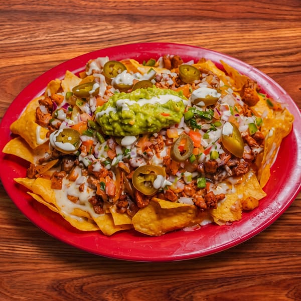 A vibrant red plate of loaded nachos with guacamole, jalapeños, and white sauce on a wooden table.