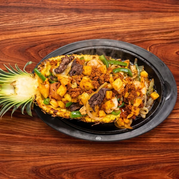 Pineapple half filled with steak, chorizo, shrimp, bell peppers, and onions on a black plate.