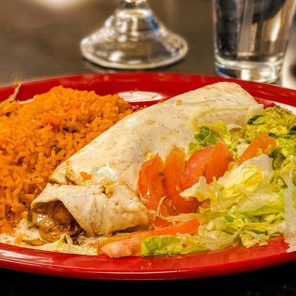 A large burrito covered in white sauce, with a side of orange rice, guacamole, lettuce, and tomatoes.