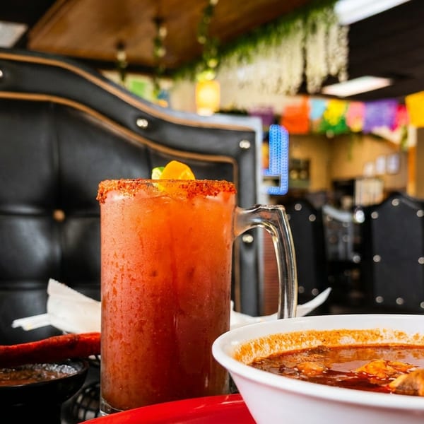 A large michelada and a bowl of menudo on a table in a vibrant Mexican restaurant.